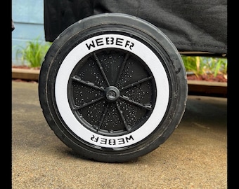 May include: Close-up of a black Weber grill wheel with a white rim. The word "WEBER" is printed in white on the rim. The wheel is on a gray surface.