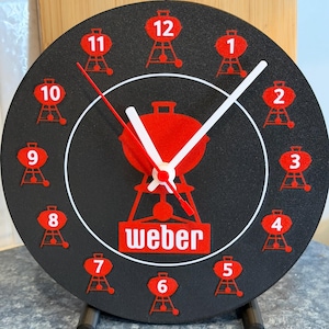 May include: A black round clock with red accents and a stand. The clock face features red grill illustrations marking the hours, with the word "Weber" in a red rectangle. The hands are white and red.