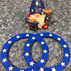 May include: A patriotic gnome figurine with a blue hat adorned with white stars, red clothing, and a white beard, grilling sausages. Two blue coasters with white stars are in the foreground. The items are themed for a holiday.