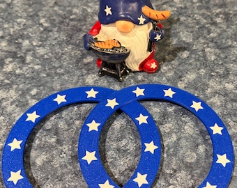 May include: A patriotic gnome figurine with a blue hat adorned with white stars, red clothing, and a white beard, grilling sausages. Two blue coasters with white stars are in the foreground. The items are themed for a holiday.