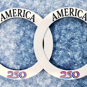 May include: Two white interlocking circular frames with the word "AMERICA" in black script at the top of each. The number "250" is printed in red, white, and blue at the bottom of each frame. The background is a textured blue surface.