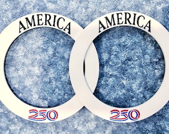 May include: Two white interlocking circular frames with the word "AMERICA" in black script at the top of each. The number "250" is printed in red, white, and blue at the bottom of each frame. The background is a textured blue surface.