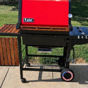 K&ouml;nnte beinhalten: Ein roter und schwarzer Weber-Grill mit einem h&ouml;lzernen Beistelltisch und einer schwarzen Kochfl&auml;che. Der Grill hat ein Thermometer und ein Rad mit patriotischem Design. Ideal zum Kochen im Freien.