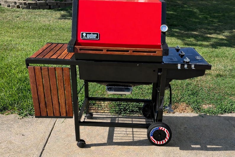 K&ouml;nnte beinhalten: Ein roter und schwarzer Weber-Grill mit einem h&ouml;lzernen Beistelltisch und einer schwarzen Kochfl&auml;che. Der Grill hat ein Thermometer und ein Rad mit patriotischem Design. Ideal zum Kochen im Freien.