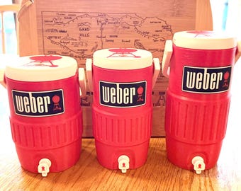 May include: Three red Weber coolers with white lids and handles are displayed. Each cooler has a white spigot and a blue and white Weber logo. A wooden box with a map design is in the background.