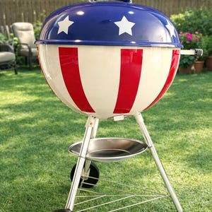 May include: A red, white, and blue outdoor grill with a domed lid featuring white stars on a blue background. The grill has a patriotic design with red stripes and a chrome-colored base and wheels. Perfect for outdoor cooking.