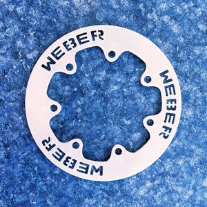 May include: A white, circular metal object with the word "WEBER" cut out in a repeating pattern. The object has several small, evenly spaced holes around its perimeter. The background is a textured, blue surface.