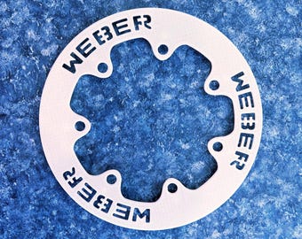 May include: A white, circular metal object with the word "WEBER" cut out in a repeating pattern. The object has several small, evenly spaced holes around its perimeter. The background is a textured, blue surface.