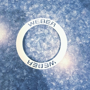 May include: A white metal ring with the word "WEBER" embossed on it twice, once on the top and once on the bottom. The ring is on a blue and white textured surface.