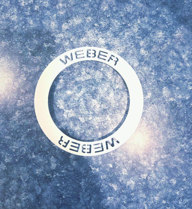 May include: A white metal ring with the word "WEBER" embossed on it twice, once on the top and once on the bottom. The ring is on a blue and white textured surface.
