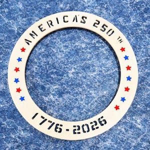 May include: A cream-colored, circular decorative item with the text "AMERICA'S 250TH" and "1776-2026" cut out. Red and blue stars are evenly spaced around the inner edge of the circle. The item is set against a blue textured background.