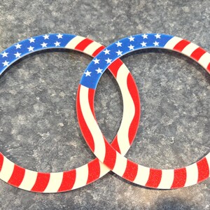 K&ouml;nnte beinhalten: Zwei ineinandergreifende kreisf&ouml;rmige Dekorationen mit patriotischem Thema. Jeder Ring hat einen blauen Abschnitt mit wei&szlig;en Sternen, der in rote und wei&szlig;e Streifen &uuml;bergeht, die der amerikanischen Flagge &auml;hneln. Das Design ist ein lebendiger Ausdruck von Nationalstolz.
