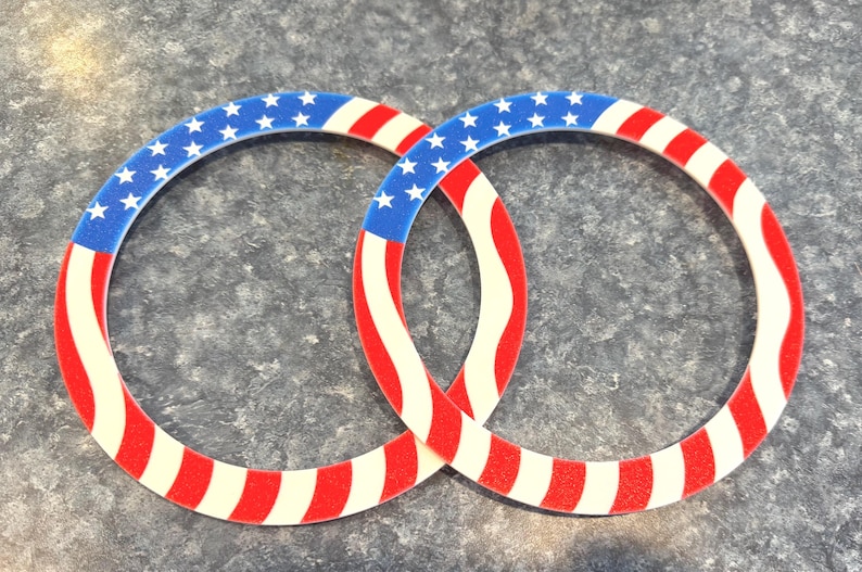 K&ouml;nnte beinhalten: Zwei ineinandergreifende kreisf&ouml;rmige Dekorationen mit patriotischem Thema. Jeder Ring hat einen blauen Abschnitt mit wei&szlig;en Sternen, der in rote und wei&szlig;e Streifen &uuml;bergeht, die der amerikanischen Flagge &auml;hneln. Das Design ist ein lebendiger Ausdruck von Nationalstolz.