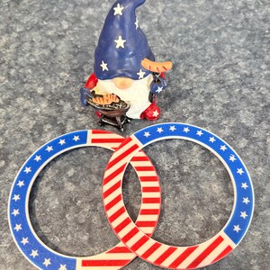 May include: A patriotic gnome figurine with a blue hat adorned with white stars, holding a sausage and a miniature grill. Two interlocking rings feature a red and white striped design and a blue border with white stars, evoking an American flag theme.