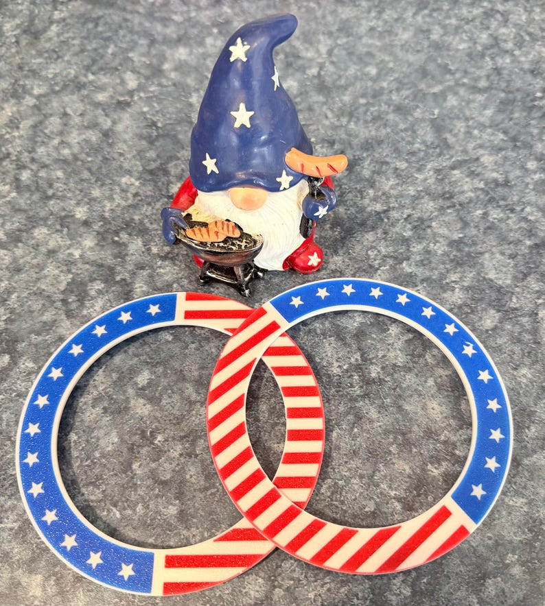 May include: A patriotic gnome figurine with a blue hat adorned with white stars, holding a sausage and a miniature grill. Two interlocking rings feature a red and white striped design and a blue border with white stars, evoking an American flag theme.