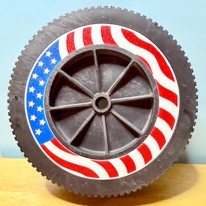 May include: A black lawnmower wheel with a tire and a central hub. The wheel features a patriotic design with red and white stripes and a blue section with white stars, resembling the American flag.