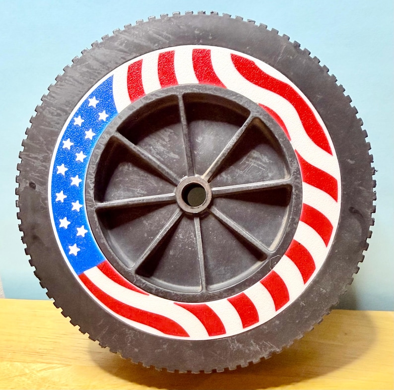 May include: A black lawnmower wheel with a tire and a central hub. The wheel features a patriotic design with red and white stripes and a blue section with white stars, resembling the American flag.