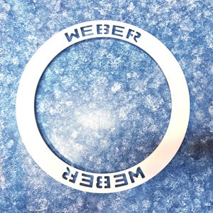 May include: A circular, flat, silver-colored metal ring featuring the word WEBER cut out twice in a bold, block-style font. The text is positioned at the top and bottom of the ring, with the bottom text oriented upside down relative to the top.