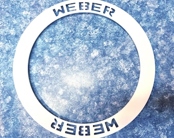 May include: A circular, flat, silver-colored metal ring featuring the word WEBER cut out twice in a bold, block-style font. The text is positioned at the top and bottom of the ring, with the bottom text oriented upside down relative to the top.