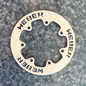 May include: A white metal ring with the word "WEBER" repeated three times around the circumference. The ring has six evenly spaced holes around the inner edge.