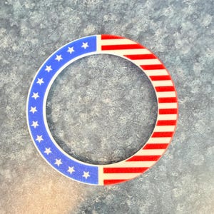 May include: Circular decorative item with a patriotic design. The item features a blue section with white stars and a red and white striped section, resembling the American flag. The design is set against a textured, blue-gray background.