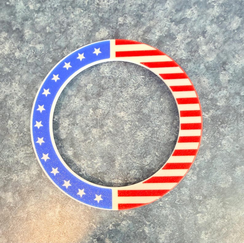 May include: Circular decorative item with a patriotic design. The item features a blue section with white stars and a red and white striped section, resembling the American flag. The design is set against a textured, blue-gray background.