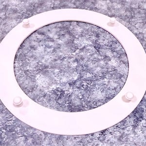 May include: A light pink, circular object with four small, rounded protrusions evenly spaced around its circumference. The object appears to be made of a solid material and is set against a textured, gray background.