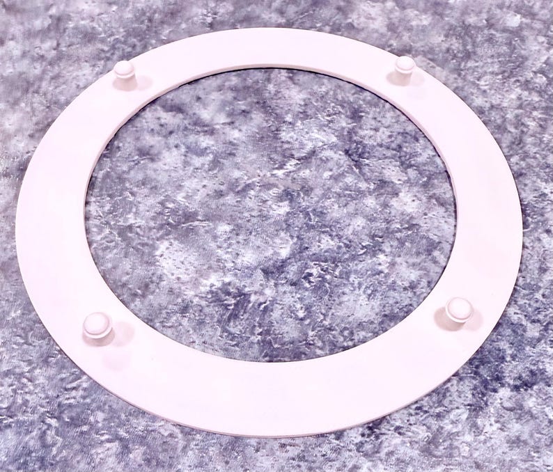 May include: A light pink, circular object with four small, rounded protrusions evenly spaced around its circumference. The object appears to be made of a solid material and is set against a textured, gray background.