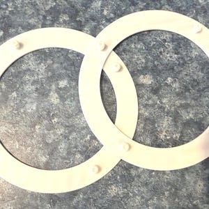 May include: Two interlocking white plastic rings with small, evenly spaced pegs. The rings are positioned on a mottled gray and blue surface, creating a simple, geometric design. The pegs appear to be for securing the rings together.