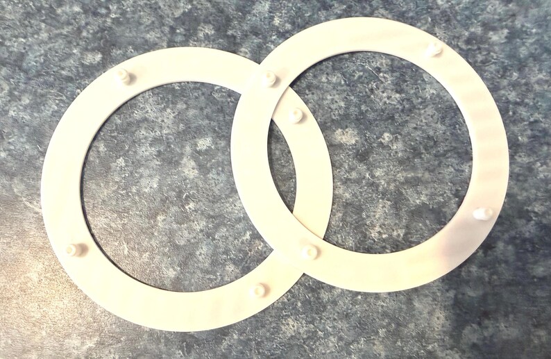 May include: Two interlocking white plastic rings with small, evenly spaced pegs. The rings are positioned on a mottled gray and blue surface, creating a simple, geometric design. The pegs appear to be for securing the rings together.