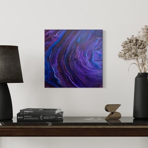 Könnte beinhalten: Quadratisches abstraktes Kunstwerk mit einem Wirbelmuster in Lila, Blau und Rot. Eine schwarze Lampe mit grauem Schirm, eine schwarze Vase mit Trockenblumen und Bücher stehen auf einem dunklen Holztisch.