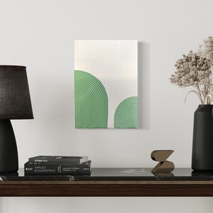 Könnte beinhalten: Abstraktes Gemälde mit strukturiertem weißem Hintergrund und zwei grünen Bogenformen. Das Kunstwerk hängt über einem dunklen Holztisch mit einer schwarzen Lampe, Büchern und einer Vase mit getrockneten Blumen.