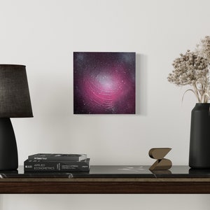 Op de afbeelding: Een vierkant abstract schilderij met een wervelend melkwegstelsel in roze, paars en wit hangt aan een witte muur. Een zwarte lamp met een grijze kap, een zwarte vaas met gedroogde bloemen en een donkere houten tafel zijn zichtbaar.