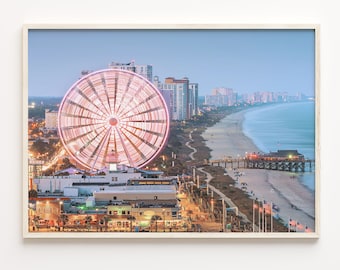 May include: A brightly lit Ferris wheel with pink and white lights stands out against a backdrop of a beach town at dusk. The Ferris wheel is located on the beach, with a pier extending out into the ocean. The beach is lined with buildings and palm trees.