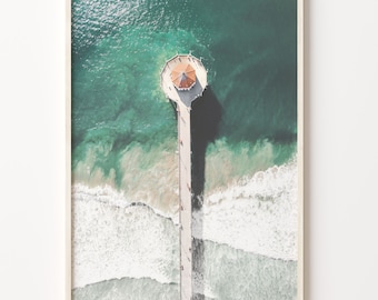 May include: An aerial view of a pier extending out into the ocean. The pier has a small, octagonal building with a red roof at the end. The water is a vibrant turquoise color, and white waves are crashing around the pier.