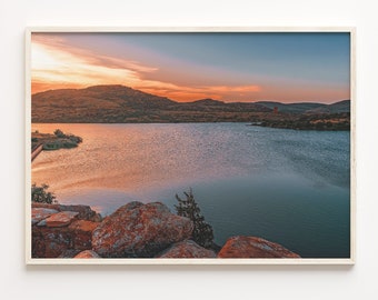 Peut inclure: Une photographie de paysage représentant un lac calme avec une chaîne de montagnes en arrière-plan. Le ciel est d'un orange et d'un bleu vifs, avec des nuages qui se reflètent dans l'eau. Au premier plan, on voit de gros rochers et un petit buisson.