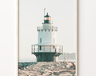 May include: A white lighthouse with a black roof and a black railing stands on a rocky shore. The lighthouse is surrounded by water and the sky is a light blue.