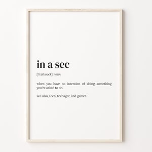 May include: A framed print with the phrase "in a sec" in a black, minimalist font. The definition of the phrase is included below the title, along with related terms. The frame is a light wood color.