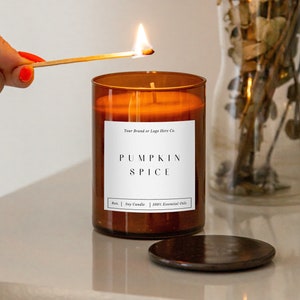 May include: A lit candle in a brown glass jar with a white label that reads "PUMPKIN SPICE". The candle is 8 oz. and made with soy wax and 100% essential oils. A match is lit above the candle.