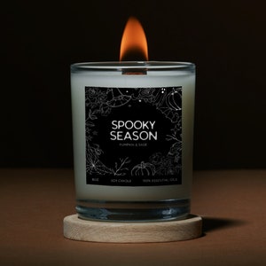 May include: A black and white illustrated candle label with the text "Spooky Season Pumpkin & Sage" and the words "Soy Candle" and "100% Essential Oils". The candle is in a clear glass jar with a wooden lid and a lit wick.