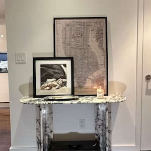 Puede incluir: Una consola de mármol blanco con una impresión enmarcada en negro de un paisaje y un mapa enmarcado en negro de la ciudad de Nueva York. Una vela blanca con el texto "New York City" está sobre la mesa.