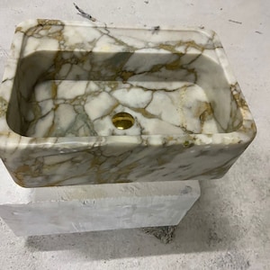 Könnte beinhalten: Ein weißes Marmorwaschbecken mit goldenen Adern. Das Waschbecken hat eine rechteckige Form und ein einzelnes Abflussloch. Das Waschbecken steht auf einem weißen Marmorsockel.