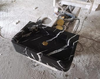 May include: A black marble sink with white veining. The sink has a rectangular basin with rounded corners and a curved front. A gold-colored drain cover is visible. The sink is on a concrete surface.