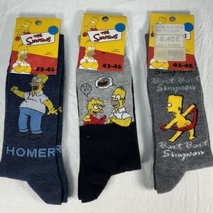 May include: Three pairs of The Simpsons character socks. One pair is navy blue with Homer Simpson, another is black and grey with Lisa and Homer, and the third is grey with Bart Simpson. Each pair has a yellow tag.
