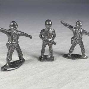 Puede incluir: Tres soldados de juguete plateados en varias poses. Uno tiene los brazos extendidos, otro sostiene un rifle y el tercero tiene un brazo levantado. Cada figura está detallada con cascos y uniformes. Juguete clásico.