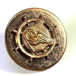 May include: A round, antique-style button with a gold-colored emblem featuring a winged design within a ship's wheel. The button has a distressed, aged appearance with a bronze-colored outer ring.