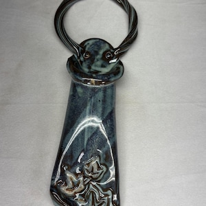 May include: A ceramic wall hanging featuring a glossy, dark blue and grey marbled glaze. The piece has a twisted circular loop at the top and a rectangular body with a flared bottom edge, decorated with a raised, embossed floral pattern.