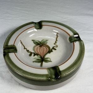 May include: Round ceramic ashtray with a floral design. The ashtray features a cream-colored interior with a brown and green border. The center has a stylized flower with green leaves and a peach-colored bloom. The ashtray has four cigarette rests.