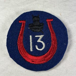 May include: A circular blue patch with a red horseshoe design and the number 13 in white. A black silhouette of an object is above the horseshoe. The patch is likely a military or historical emblem.