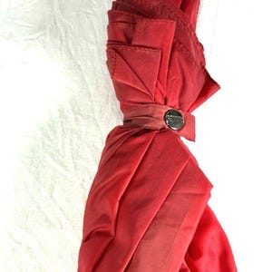May include: A vintage red umbrella featuring a wooden handle crafted in the shape of a goose's head. The goose's beak is painted red, and the umbrella is secured with a matching red fabric strap and a silver clasp.
